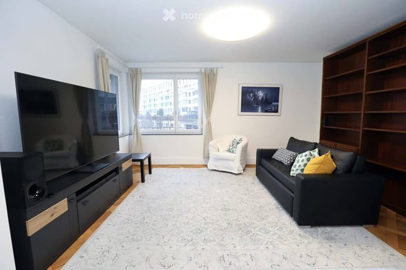 Modern living room with a large flat-screen TV on a black stand, a black sofa with colorful throw pillows, a white armchair, a coffee table, and a bookshelf with built-in storage. The room features large windows with curtains and a patterned rug on the wo