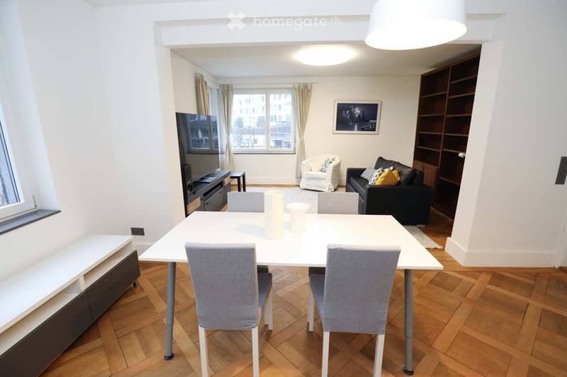 A modern, well-lit living and dining area with wooden flooring, white walls, and large windows. Features include a white dining table with four chairs, a black sofa with decorative pillows, a white armchair, a flat-screen TV on a stand, and a built-in boo