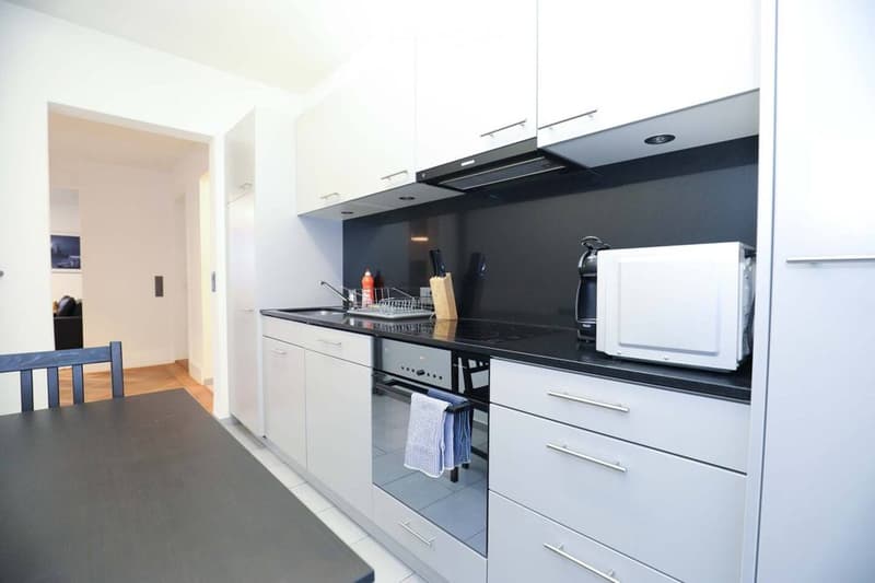 Modern kitchen with white cabinets, black countertops, stainless steel appliances, and a small dining area with a table and chair.