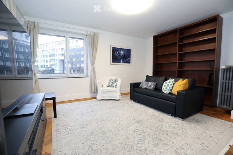 Modern living room with a black sofa, white armchair, coffee table, bookshelf, and a large window with curtains.