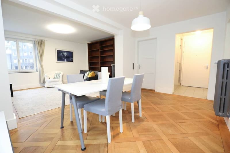 Bright dining and living area with wooden flooring, white dining table with four chairs, a small sofa with cushions, a bookshelf, and a large window with curtains.