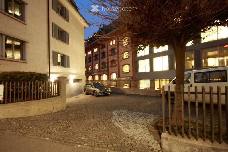 Evening view of a multi-story residential building with illuminated windows, parked cars, and a cobblestone driveway.