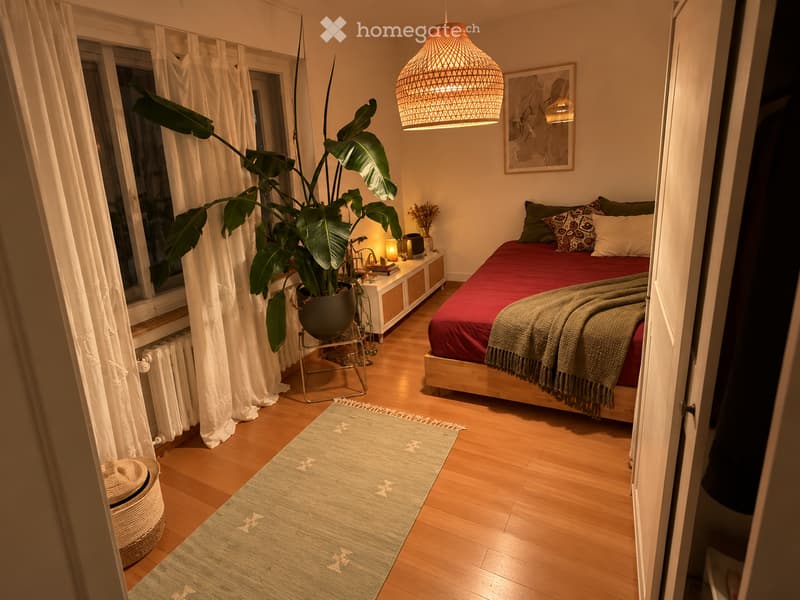 Cozy bedroom with wooden flooring, a large bed with red bedding, a nightstand with a lamp, a potted plant, and a wicker basket near the door.