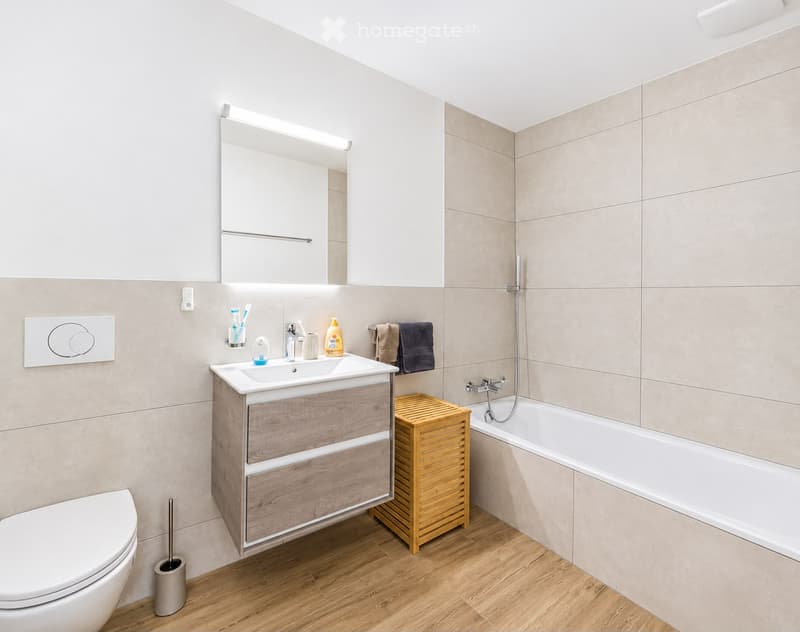 Modern bathroom with a wooden sink cabinet, white tiles, a bathtub, and a toilet. Features include a mirror with built-in lighting, a wooden storage unit, and neatly arranged towels and toiletries.