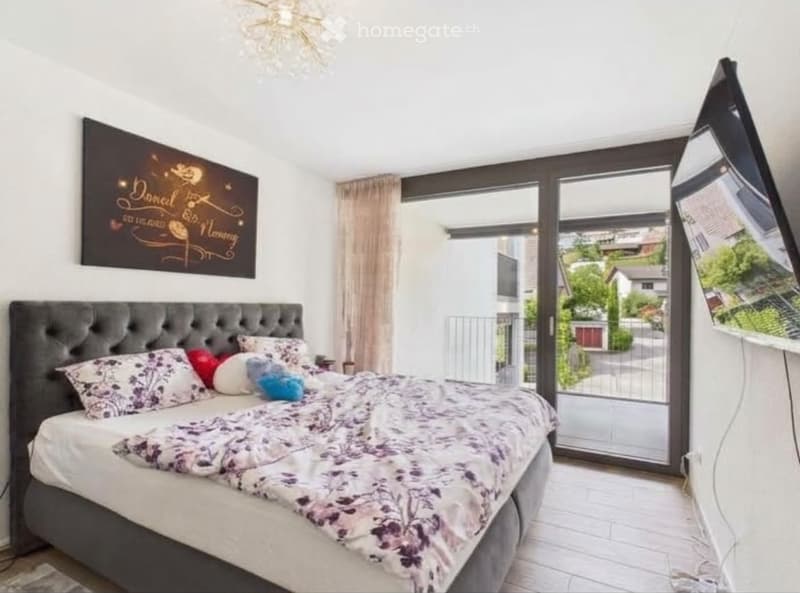 Modern bedroom with a large bed, tufted headboard, decorative pillows, and a mirror on the wall. Large windows offer a view of the outdoor area.