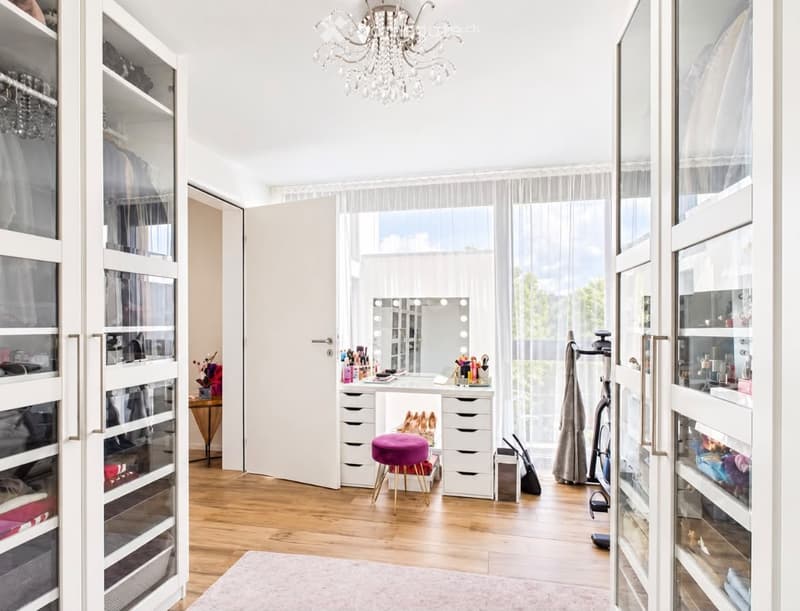 Well-lit dressing room with a vanity, large mirror, purple stool, wardrobe, and chandelier.