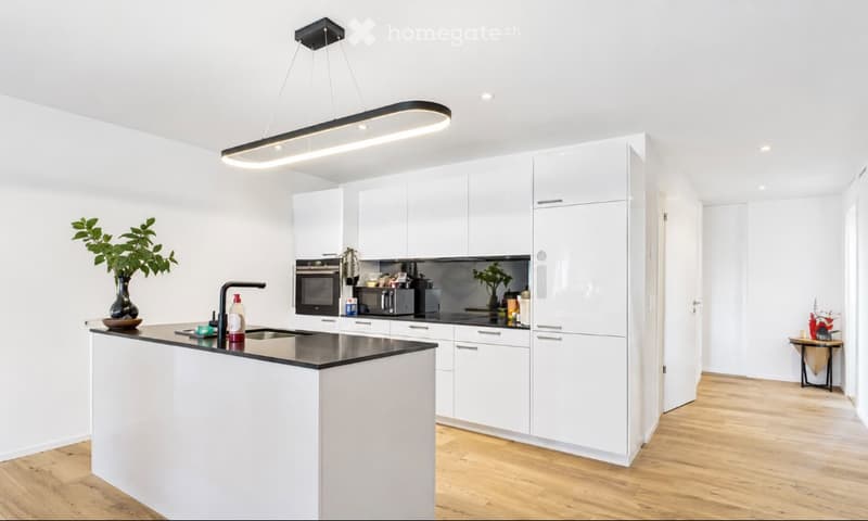 Modern kitchen with white cabinets, a central island, and various appliances including an oven and microwave. The space features a sleek, minimalist design with a pendant light fixture and a small decorative table in the corner.