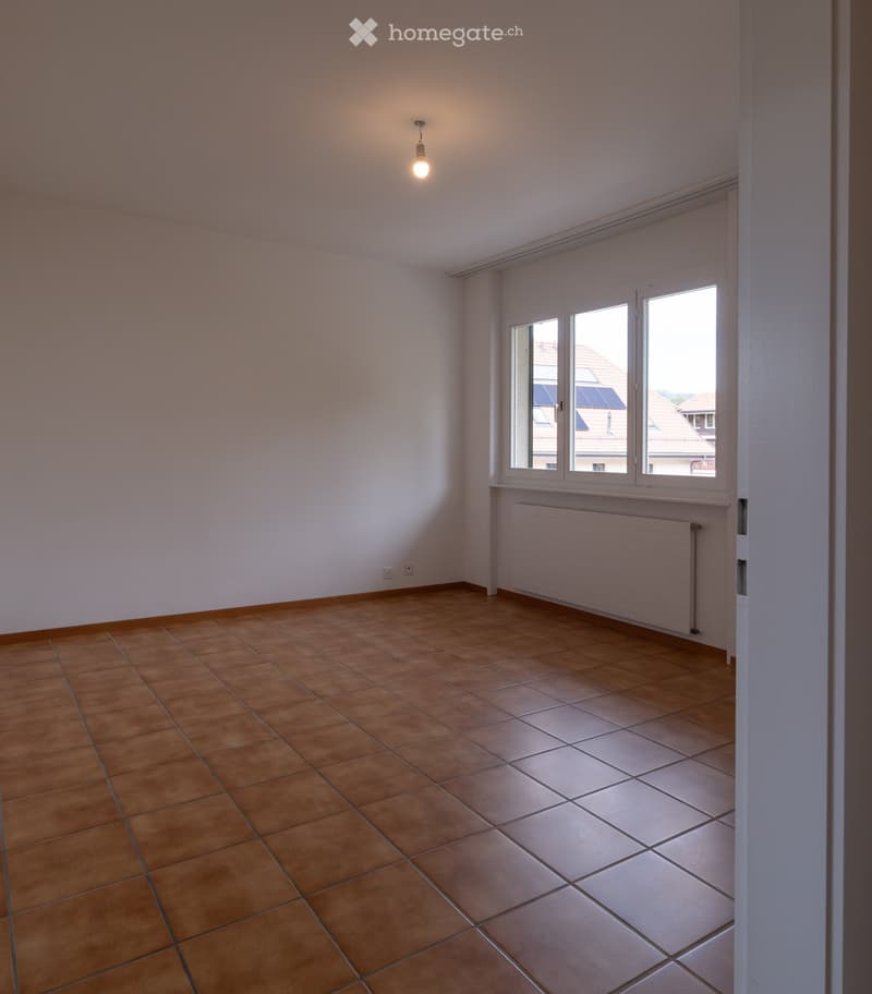 Empty room with tiled floor and large windows, featuring a radiator and a single light fixture.