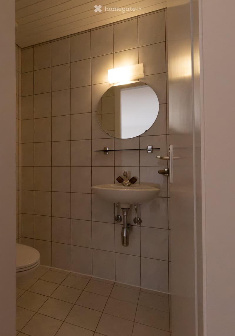 Modern bathroom with beige tiled walls and floor, a round mirror with built-in lighting, a wall-mounted sink with a chrome faucet, and a toilet visible in the background.