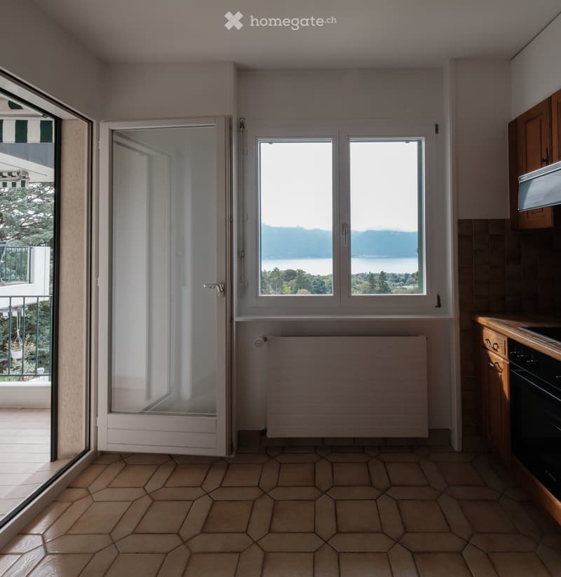 Empty kitchen with tiled floor, wooden cabinets, and large windows offering a scenic view of a lake and mountains.