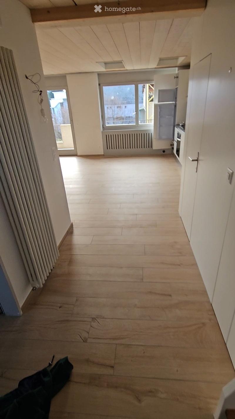 Empty room with light wooden flooring, white walls, and a radiator on the left side. A doorway leads to another room with a window and a small kitchen area visible in the background.