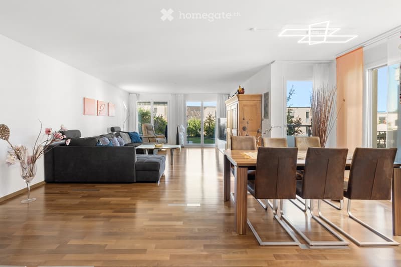 Open-plan living and dining area with wooden flooring, large windows, modern furniture including a sectional sofa, coffee table, ottoman, dining table with chairs, and decorative vases.