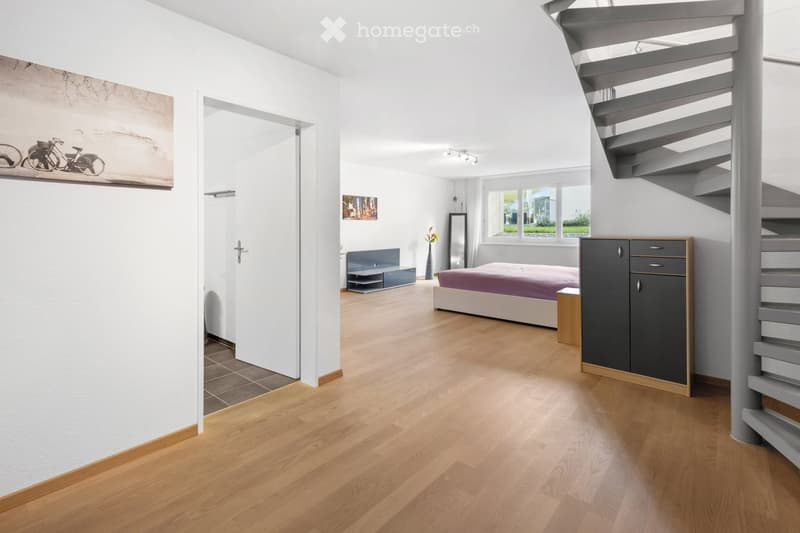 Modern bedroom with wooden flooring, a bed with a purple bedspread, a nightstand, a cabinet, a mirror, and a vase with flowers. A staircase is visible on the right side.