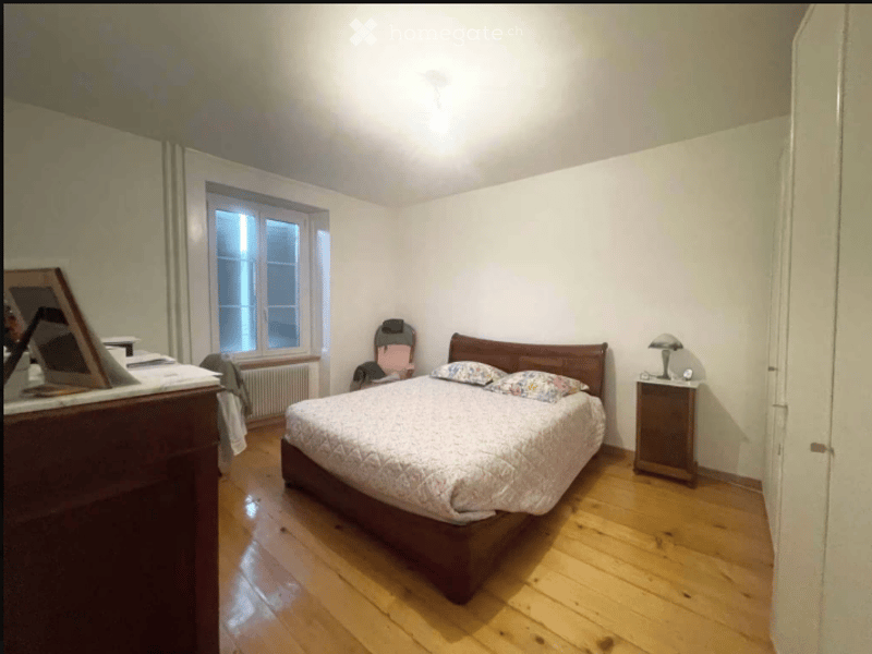 Cozy bedroom with wooden flooring, a large bed with patterned bedding, a nightstand with a lamp, a chair, and a radiator under a window.