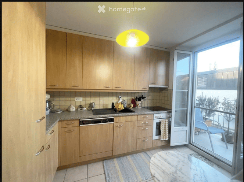 Bright kitchen with light wooden cabinets, granite countertops, and stainless steel appliances including a dishwasher and oven. A sliding glass door opens to a balcony with outdoor furniture.
