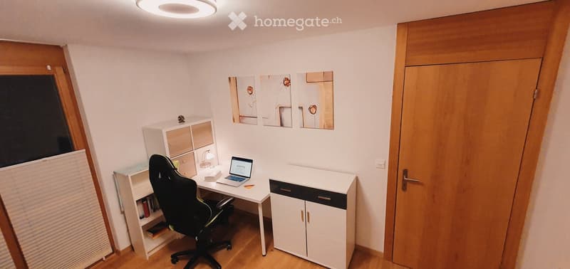A modern, well-lit room with a desk, chair, laptop, and cabinet. The room features a large window with blinds, a wooden floor, and several picture frames on the wall.