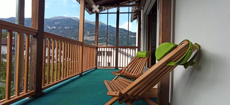 Balcony with wooden railing, green carpet, two wooden chairs, and a scenic mountain view.