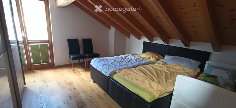 Cozy attic bedroom with wooden slanted ceilings, a double bed with yellow and blue bedding, two black chairs, and large windows letting in natural light.