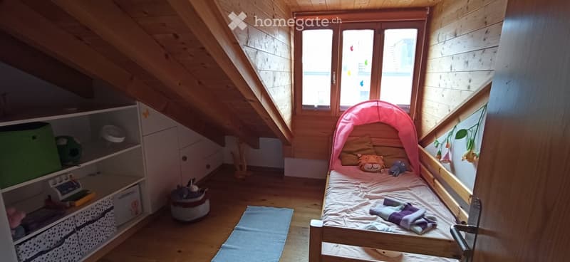 Cozy attic bedroom with wooden walls and slanted ceilings, featuring a small bed with a pink canopy, stuffed toys, a shelf with storage boxes, and a rug on the floor.