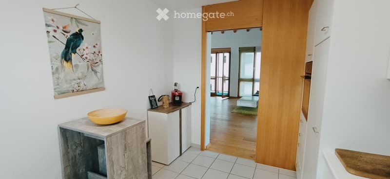 A modern hallway with white tiled flooring, wooden doors, and a small wooden table with a yellow bowl and a few decorative items. A framed artwork hangs on the wall, and the hallway leads to another room with wooden flooring and a visible piece of furnitu