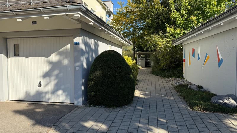 A paved pathway leads to a white garage with a large, round, trimmed bush in front. The garage has a small window and a door handle. Flags are attached to the adjacent building.
