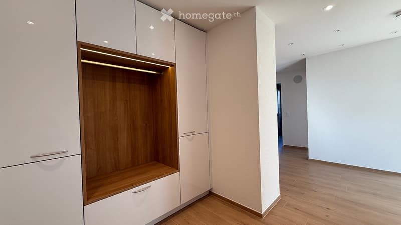 Empty hallway with wooden flooring, white walls, and built-in storage unit with wooden shelves and white cabinets.