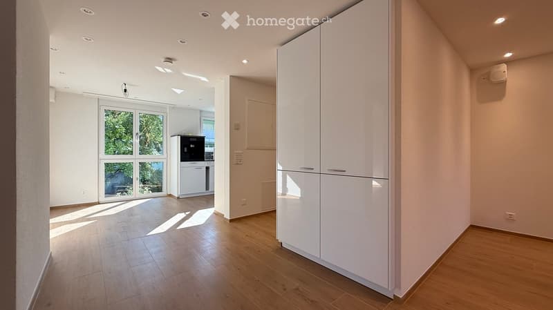 Bright, empty hallway with wooden flooring, white walls, recessed lighting, and a large window at the end leading to another room.