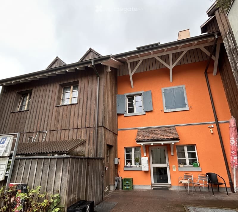 Two-story building with orange and wooden facades, multiple windows with shutters, small balcony, and outdoor seating area with chairs.
