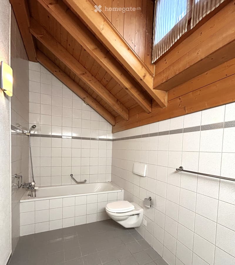 Bathroom with a bathtub, toilet, tiled walls, and a wooden slanted ceiling with a window. Includes a showerhead and handrail.