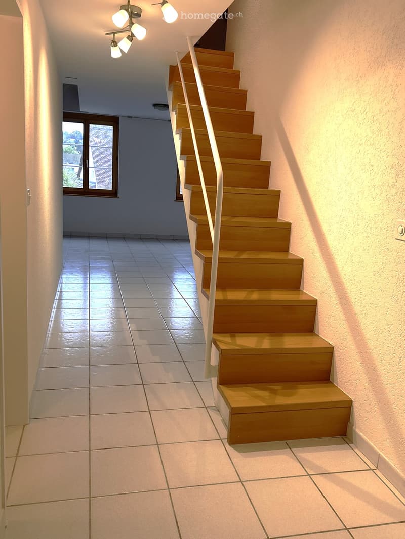 A modern staircase with wooden steps and a white handrail, illuminated by ceiling lights, leading up from a tiled hallway.