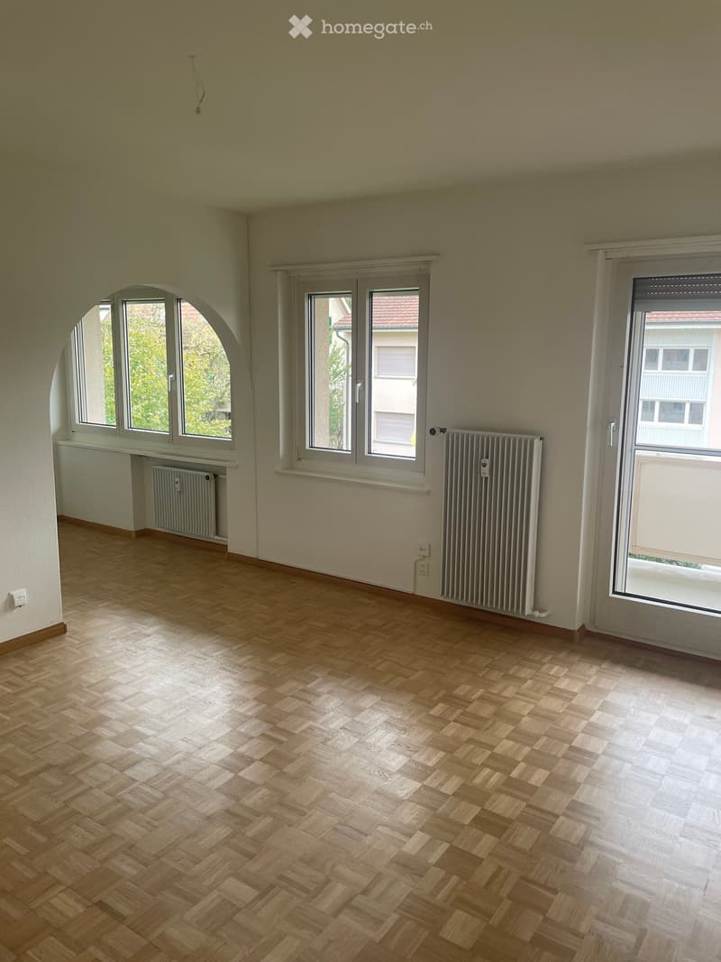 Empty room with wooden herringbone flooring, large windows, radiators, and a sliding glass door leading to a balcony.