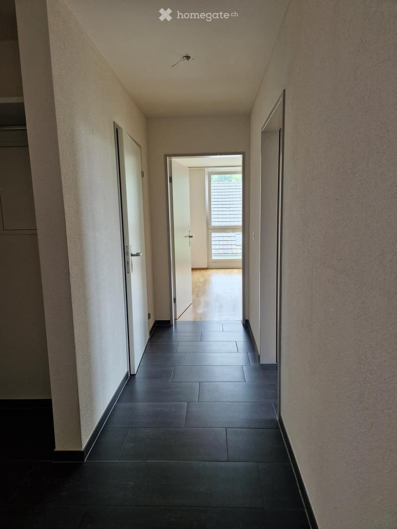 A narrow hallway with dark tiled flooring, light-colored walls, and a door leading to a room with a window.