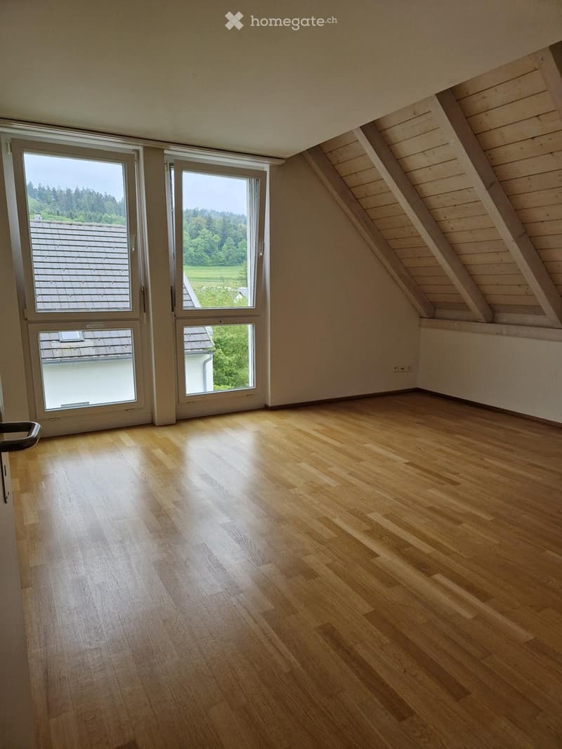 Empty attic room with wooden flooring, large windows, and slanted wooden ceiling beams.