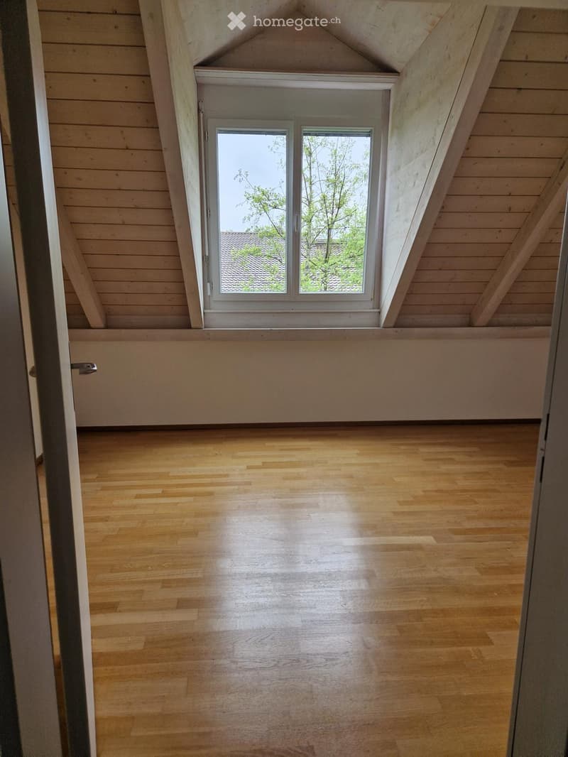 Empty attic room with wooden floors and slanted wooden ceilings, featuring a large window with white frame and glass panels.