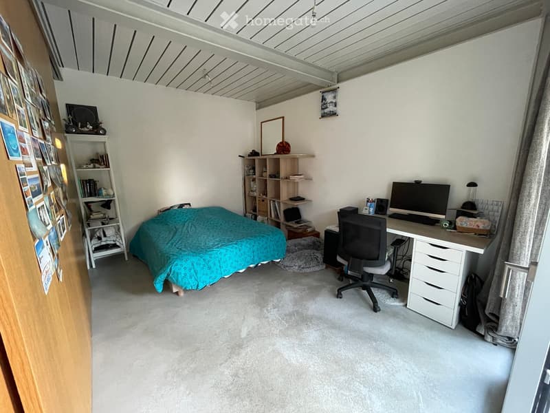 A multi-functional room with a bed, desk, and shelves. The room features a bed with a teal duvet, a desk with a computer and chair, shelves with books and decorative items, and a large window with curtains.