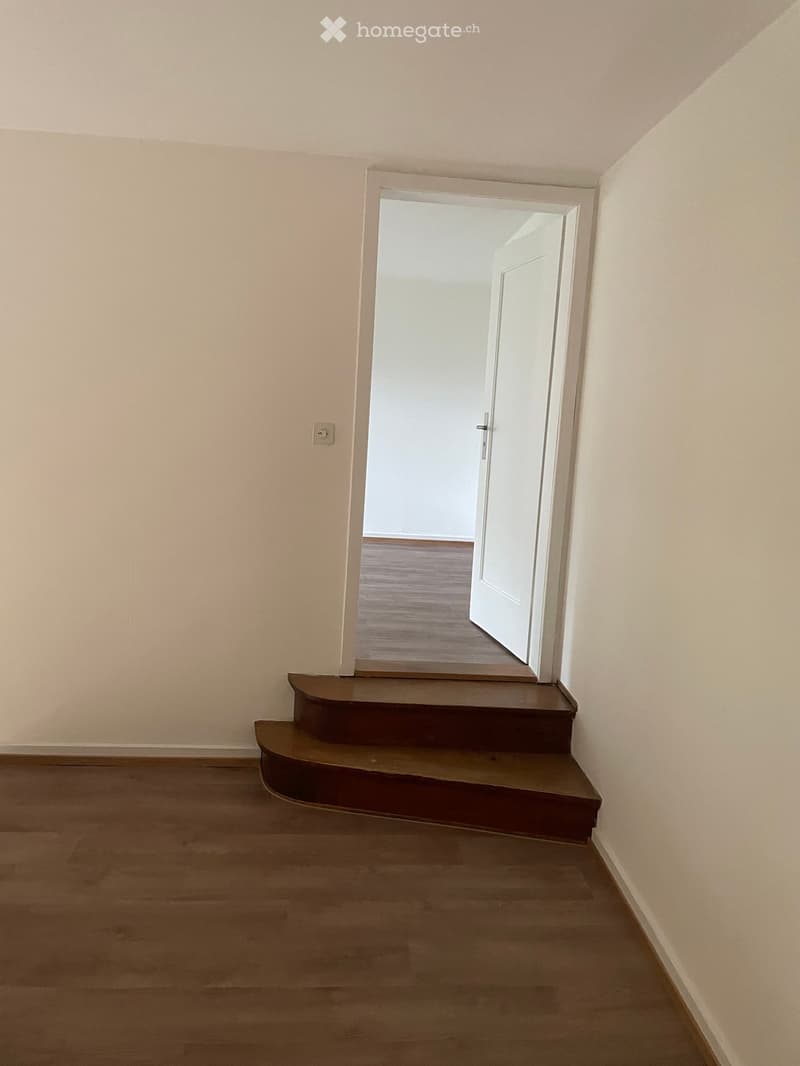 Empty room with wooden flooring, beige walls, and a small staircase leading to another room.