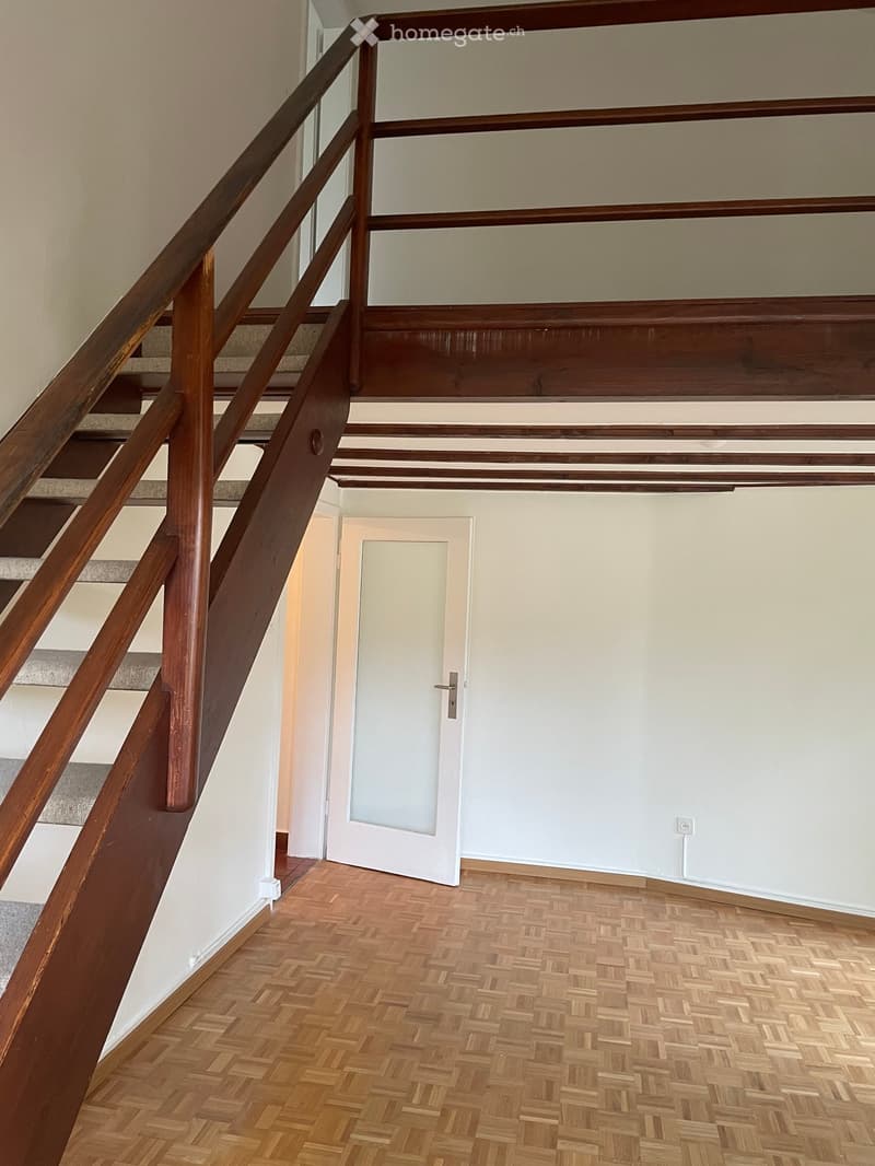 Empty hallway with wooden herringbone flooring, white walls, and a staircase with wooden railings leading upwards.