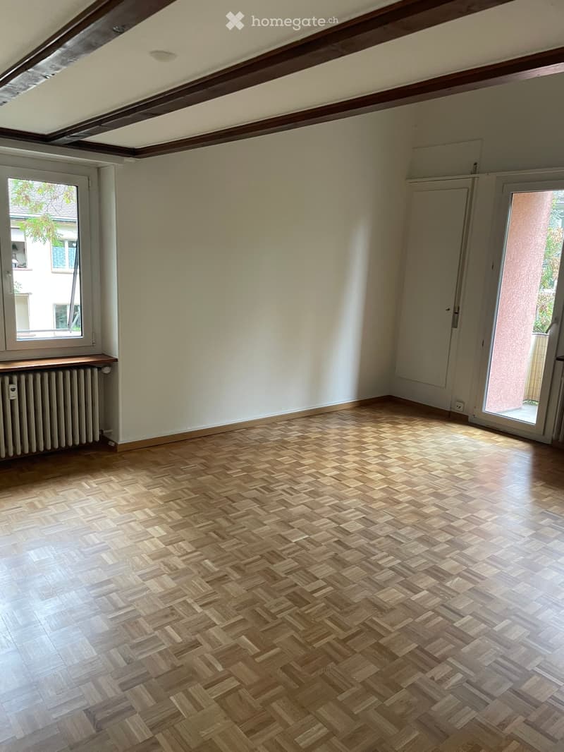Empty room with wooden herringbone flooring, white walls, large window, and sliding glass door leading outside.
