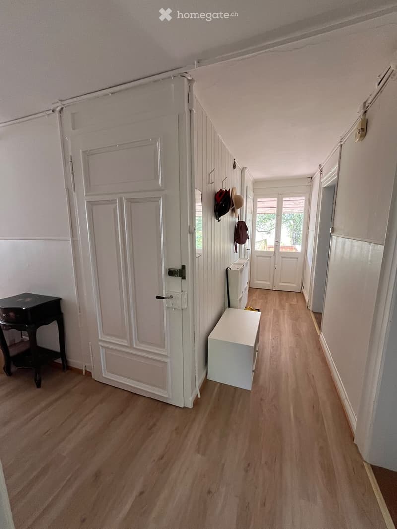 A clean, well-lit hallway with wooden flooring, white walls, and several doors. A small black table is visible on the left, and a white bench is placed near the wall on the right. A coat rack with hats and bags is mounted on the wall.