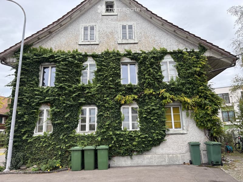 Two-story house with white exterior and green ivy covering the walls, multiple windows, and several green trash bins in front.