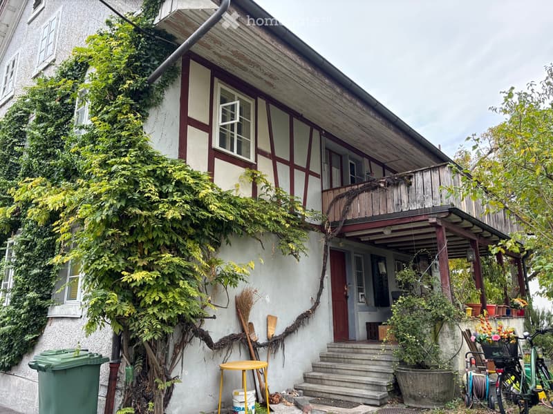 Two-story house with wooden balcony, ivy-covered walls, and small garden area with potted plants and a bicycle.