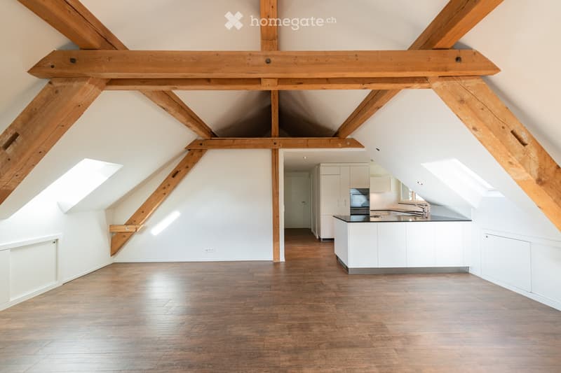 Open-plan attic space with exposed wooden beams, wooden flooring, and a modern kitchen area featuring white cabinets, a stove, and a sink.