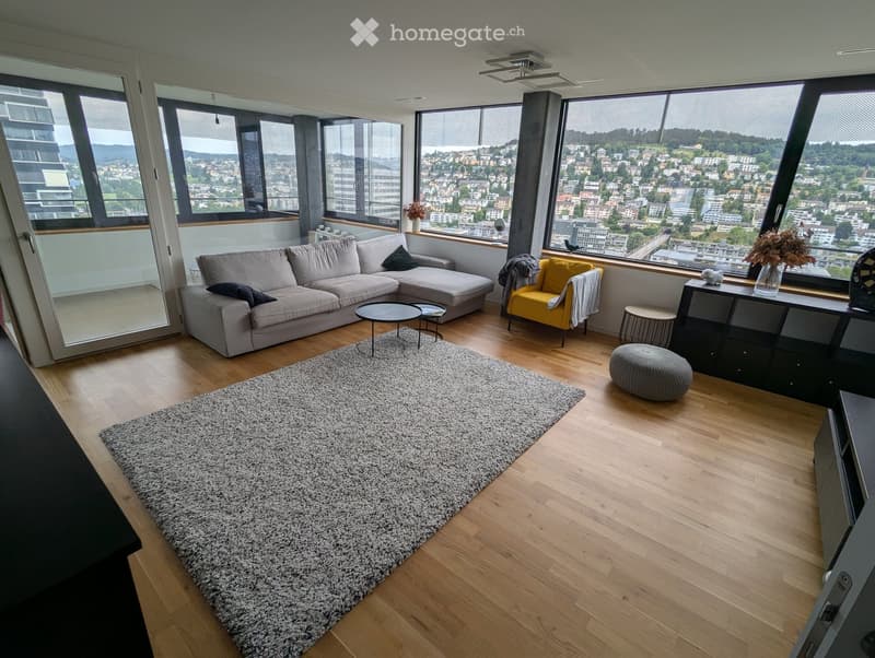 Modern living room with large windows offering a city view, featuring a grey sectional sofa, a yellow accent chair, a round coffee table, a grey ottoman, and a large shaggy rug on wooden flooring.