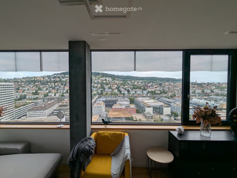 Modern furnished room with large windows offering a cityscape view, featuring a yellow sofa, a small round stool, a side table with a vase of dried flowers, and a mirror on the windowsill.