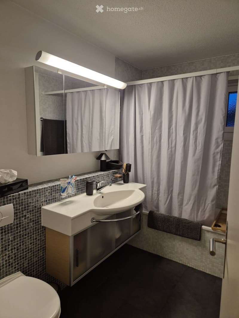 Modern bathroom with a white sink, mirror, and toilet. Features a tiled backsplash, a shower curtain, and a towel on the side.