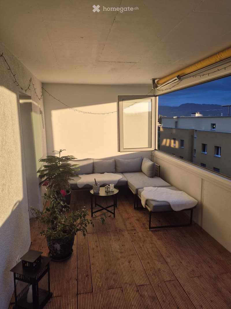 Balcony with wooden decking, grey sectional sofa, small round table, potted plants, and a view of the city and mountains.