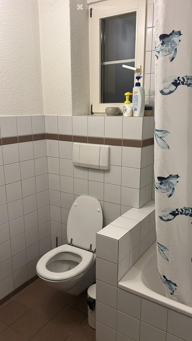 A small bathroom with white tiled walls, a toilet, a shower curtain, and a small window above the sink area.