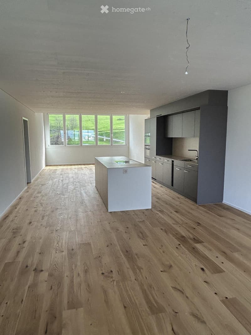 Open-plan kitchen and living area with light wood flooring, white island, grey cabinets, and large windows offering a view of greenery outside.