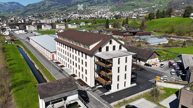 Aerial view of a modern multi-story building with a brown roof and white facade, surrounded by a parking lot, greenery, and a scenic town in the background.