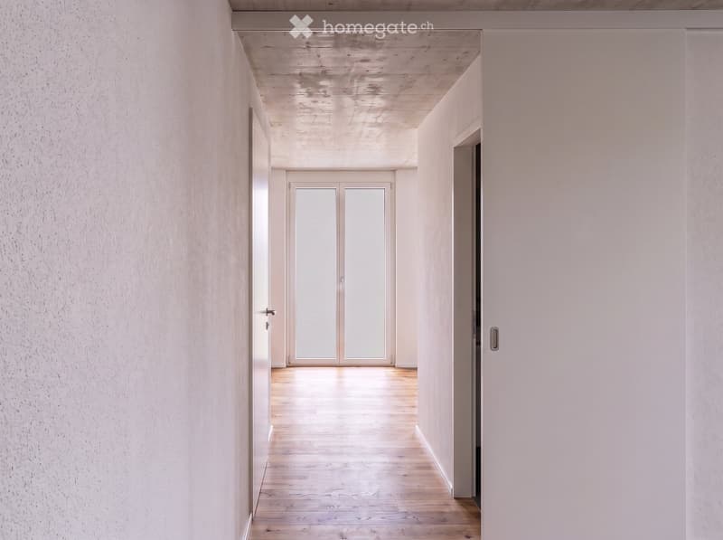 A modern, minimalist hallway with light wooden flooring, white walls, and a double glass door at the end leading to another space.